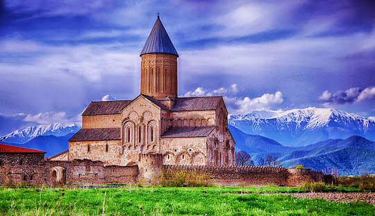 Ancient stone church with conical tower in green valley with snow-capped mountains in background, Georgia
